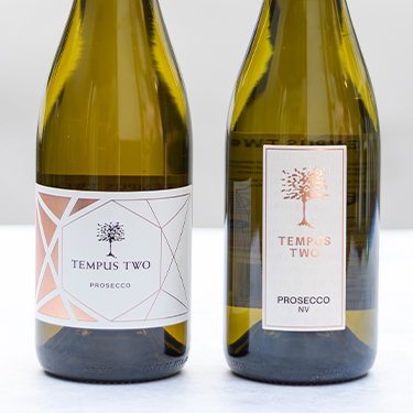 Side-by-side comparison of Tempus Two Prosecco bottles, featuring the previous geometric label design and the new minimalist label with a copper tree emblem, highlighting the brand’s packaging refresh.